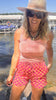 Woman in pink top and checkered shorts standing by a lake with boats in the background