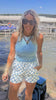 Woman in a light blue top and checkered shorts standing by a body of water with a cooler.