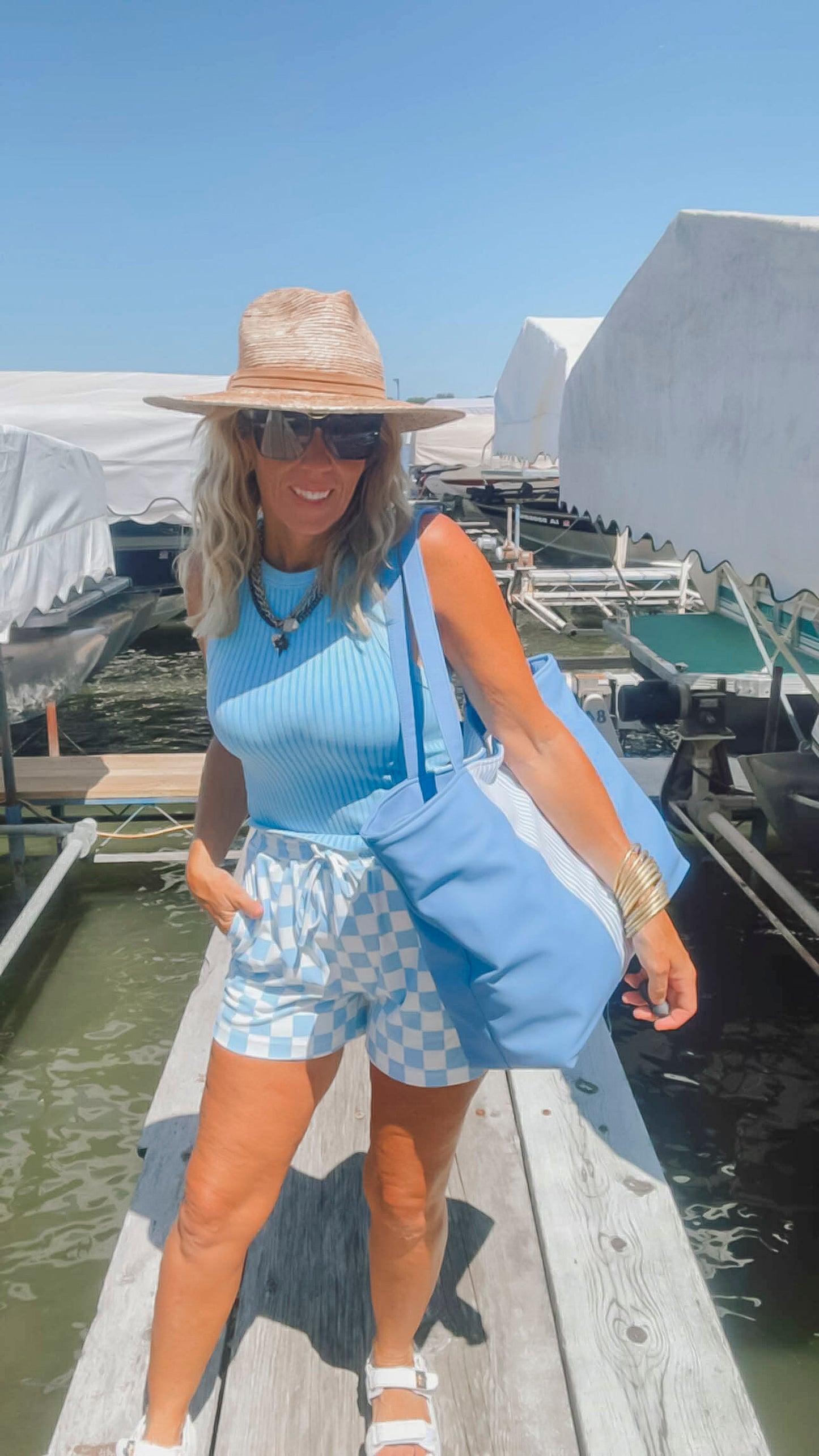 Woman on a dock wearing a blue top, checkered shorts, and a straw hat with a blue bag.