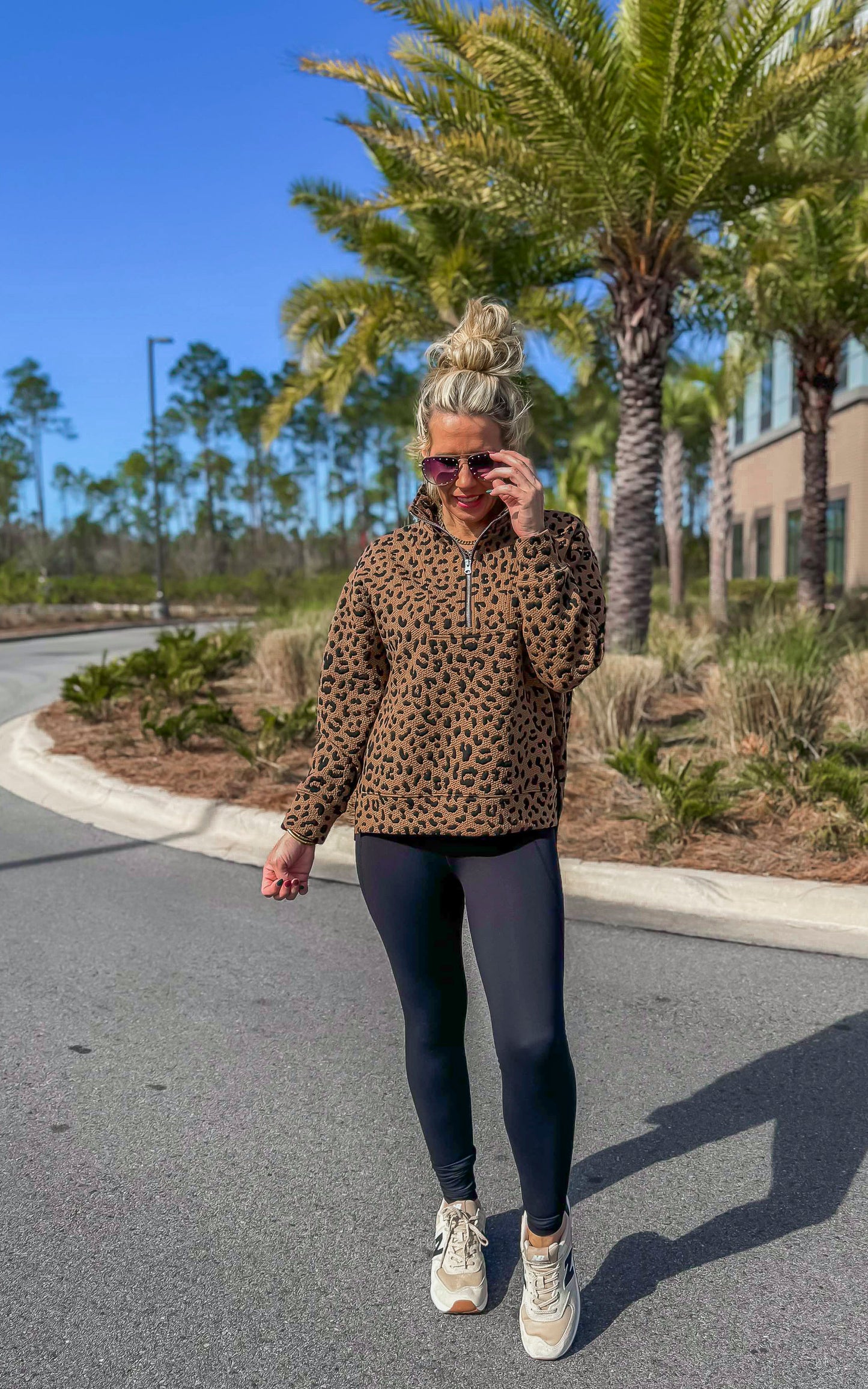 A woman standing on the road wearing a leopard print quarter zip pullover with black leggings and white sneakers.