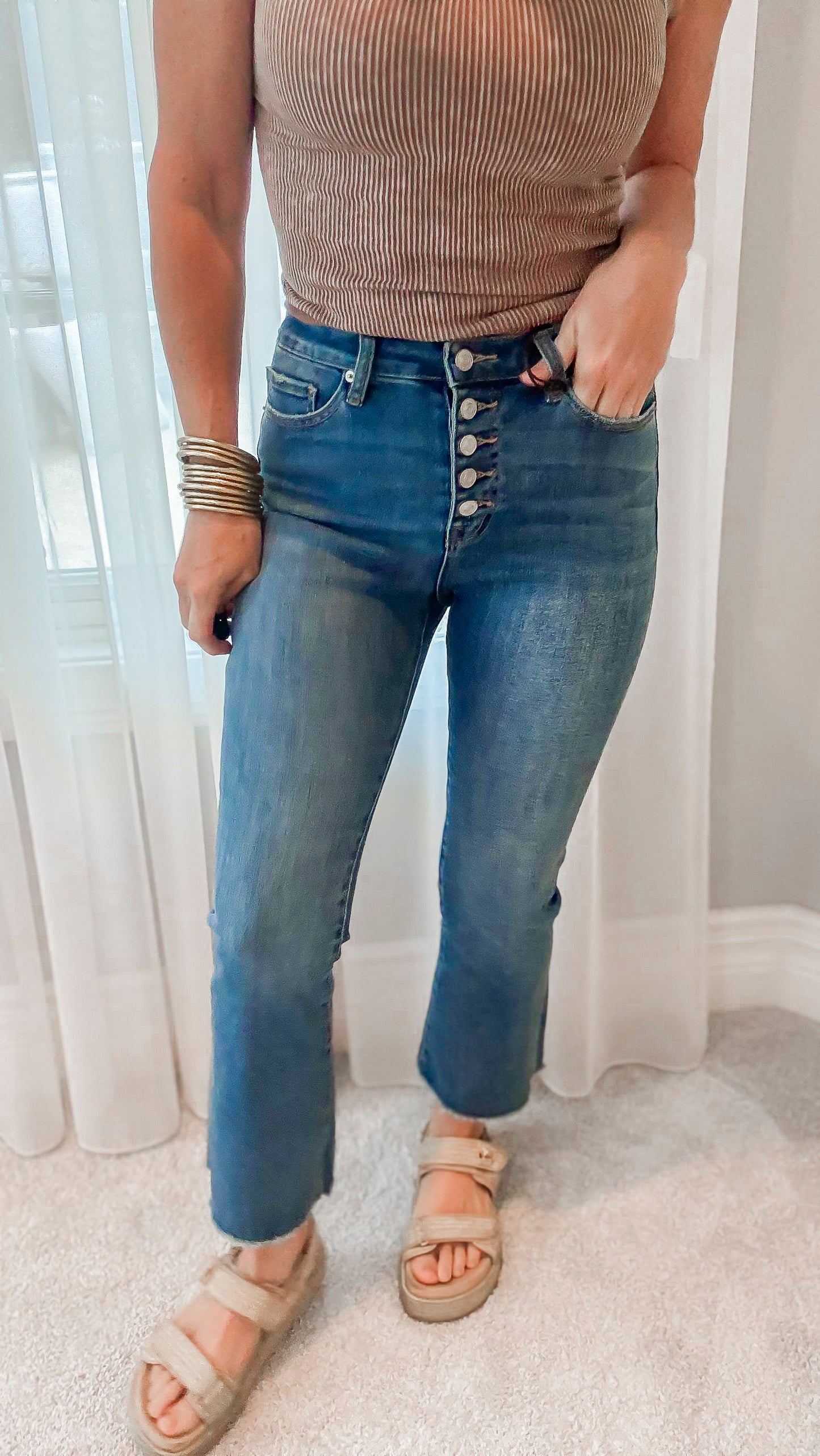 A person wearing medium blue high rise crop button up flare denim jeans and a beige top, accessorized with a belt, bracelets, and sandals.