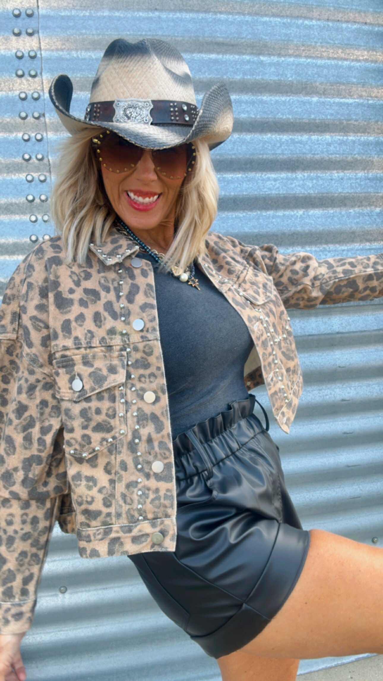 Person wearing a leopard print jacket, black top, and shorts with a cowboy hat against a corrugated metal background.