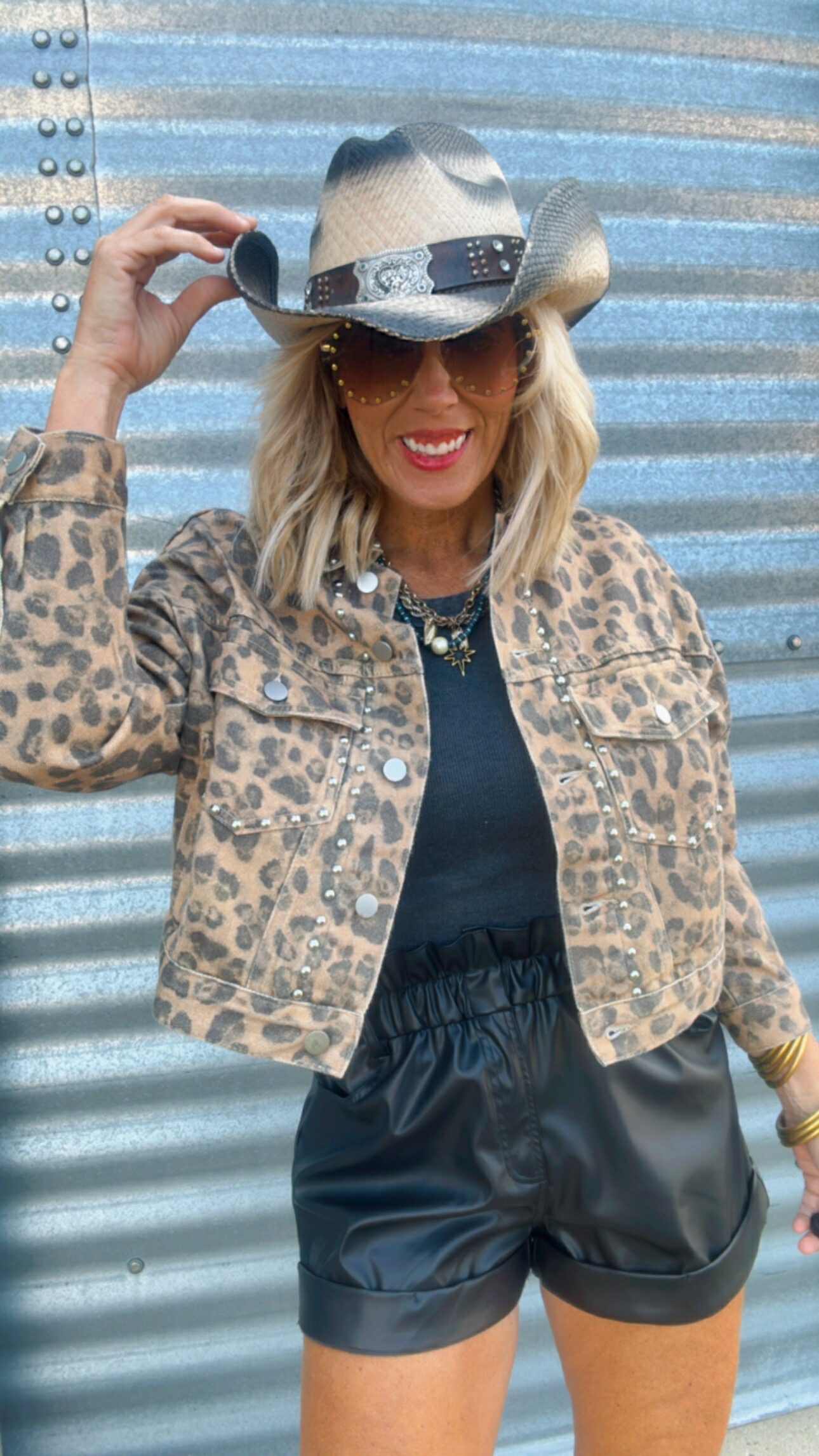 Person wearing a leopard print jacket, black shorts, and a straw hat against a corrugated metal background.