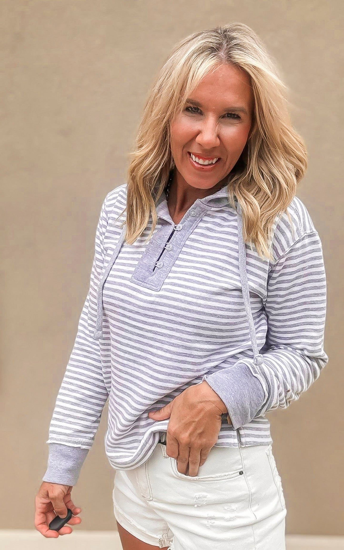 Woman wearing a grey and white striped henley hoodie with drawstrings, paired with white distressed denim shorts.