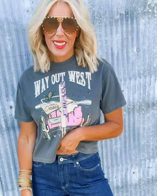 Person wearing a 'Way Out West' t-shirt with sunglasses and blue jeans against a textured light blue background