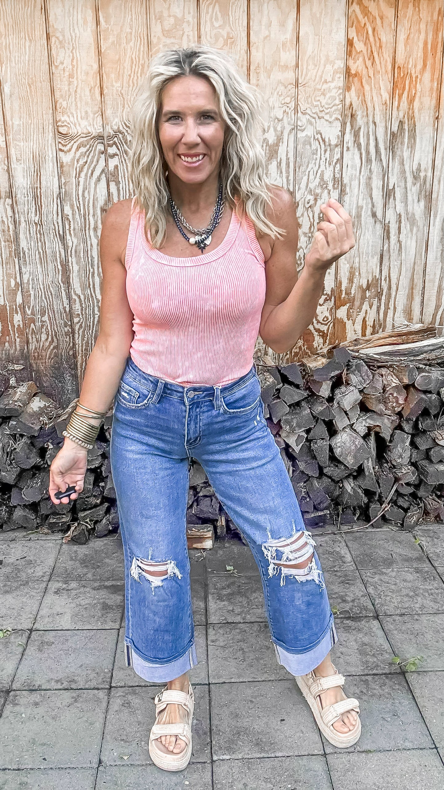 Woman wearing a pink tank top and blue jeans standing in front of a wooden wall.