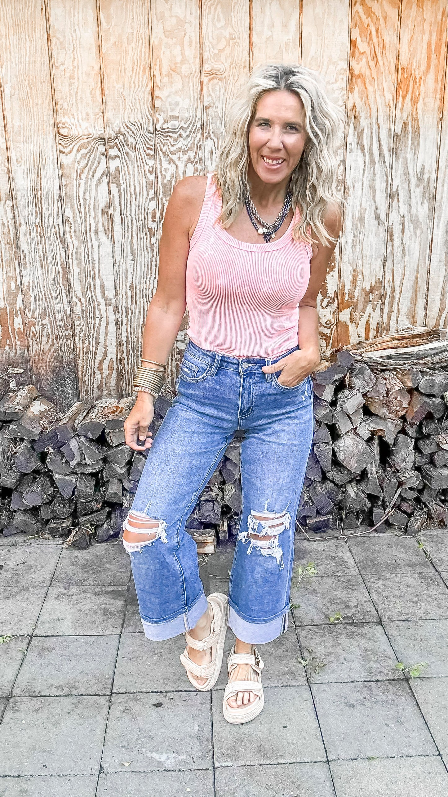 Woman wearing a pink top and ripped jeans standing in front of a wooden wall.