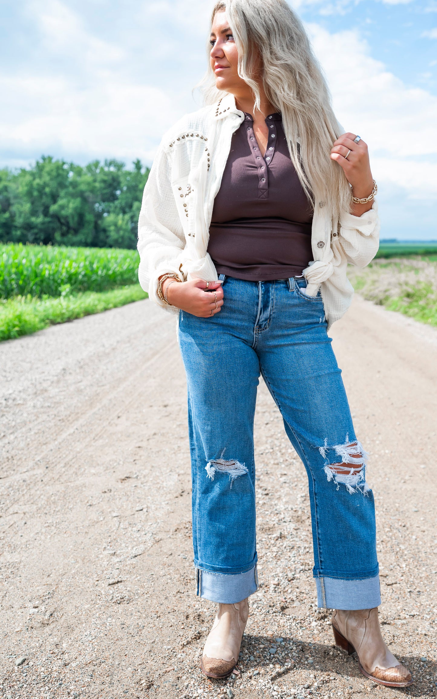 Woman standing on a dirt road wearing a white jacket, brown top, blue jeans with fish designs, and beige boots.