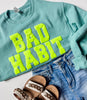 Close up of a Jerzee Sweatshirt with Neon Yellow Bad Habit on it paired with sandals and shorts.