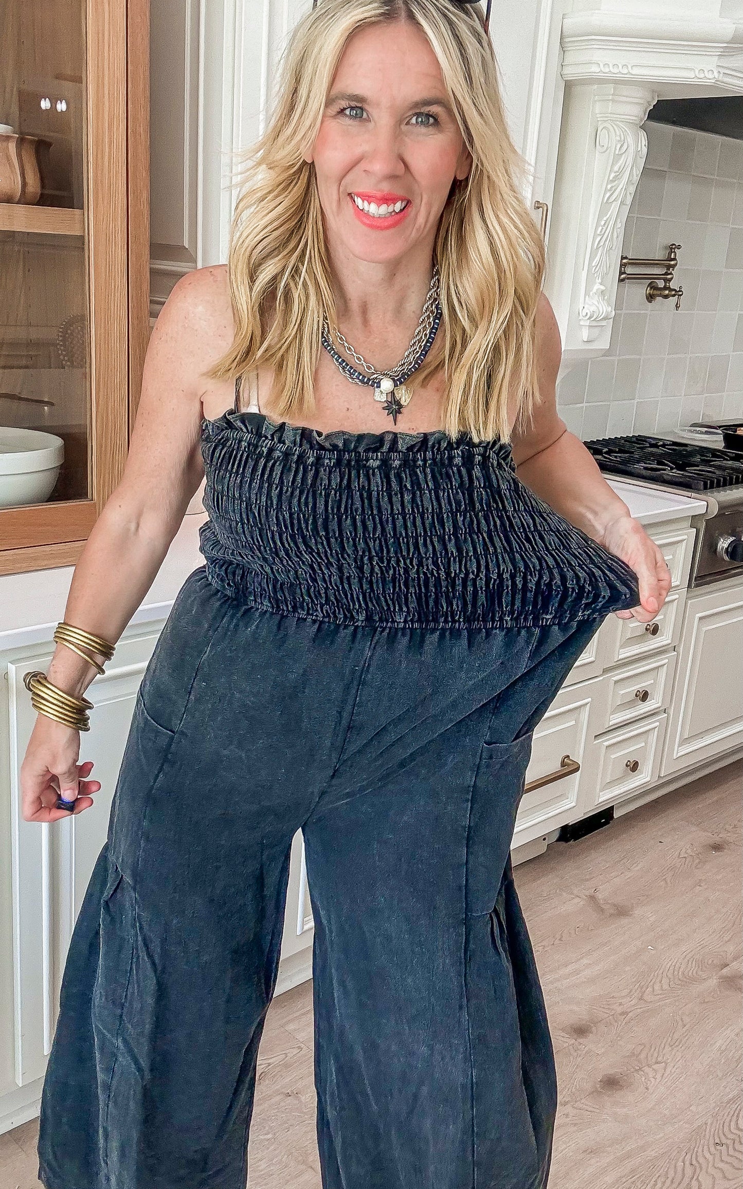 Black sleeveless jumpsuit featuring a smocked bodice and relaxed wide-leg fit, styled in a modern kitchen setting.