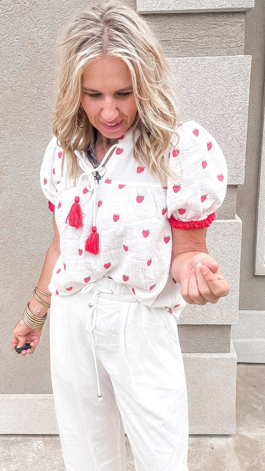 Lightweight white blouse with whimsical strawberry accents paired with casual linen trousers for a fresh, airy look.