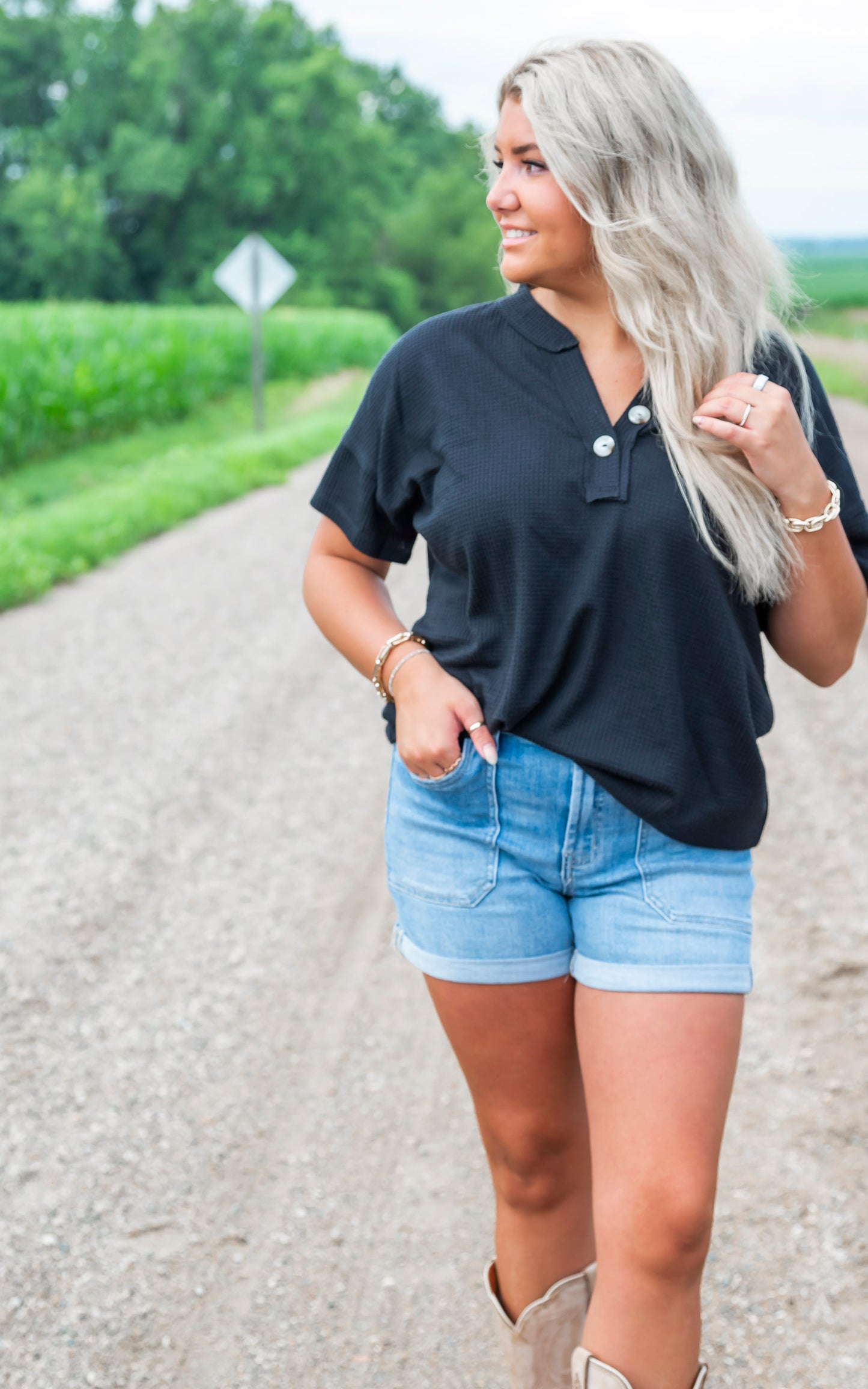Woman in black top and denim shorts standing on a gravel road with greenery in the background