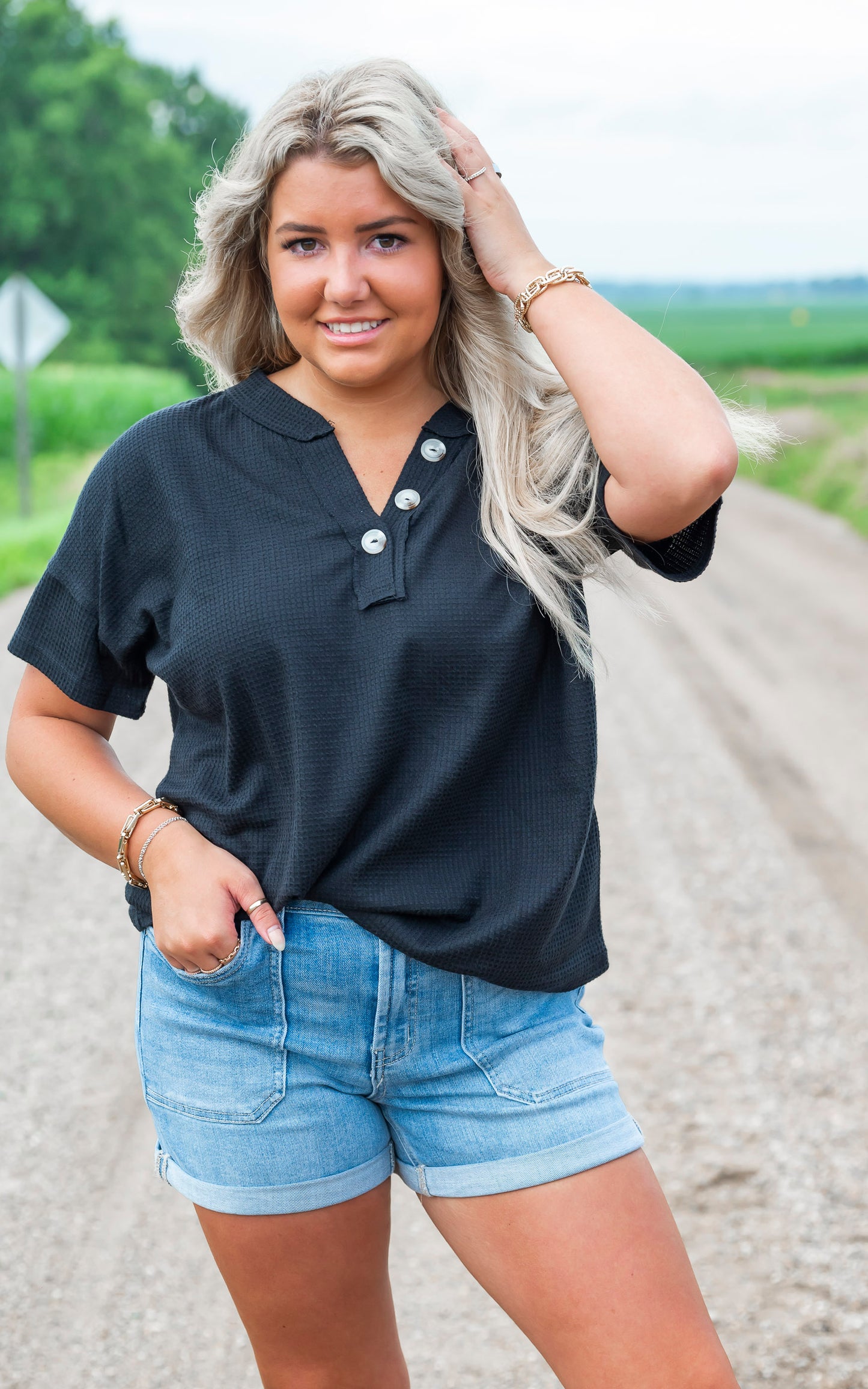 Woman wearing a black top and denim shorts standing on a gravel road with greenery in the background