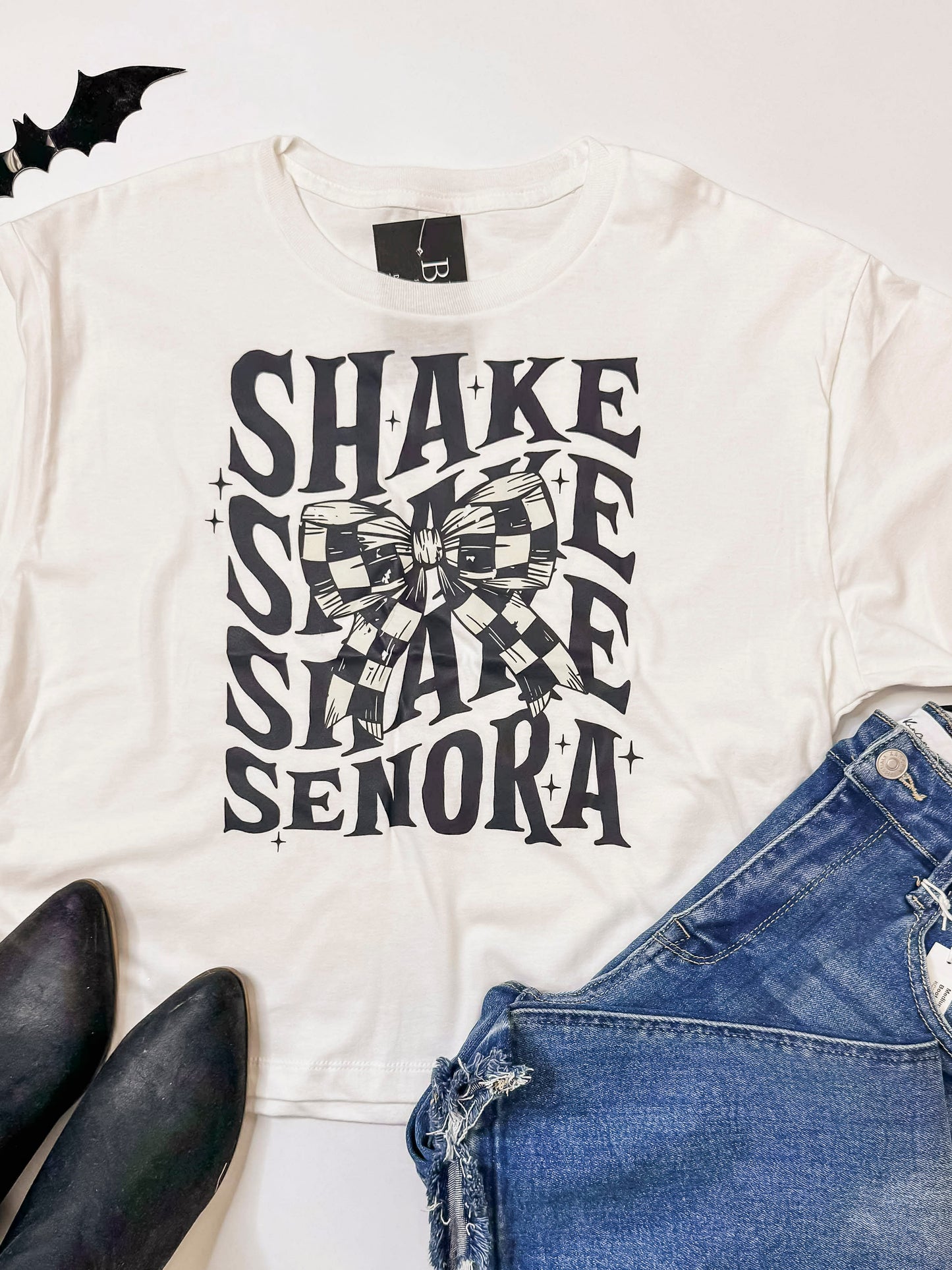 White t-shirt with 'SHAKE ME SENORA' text and bow graphic, paired with denim shorts and black shoes on a white background.