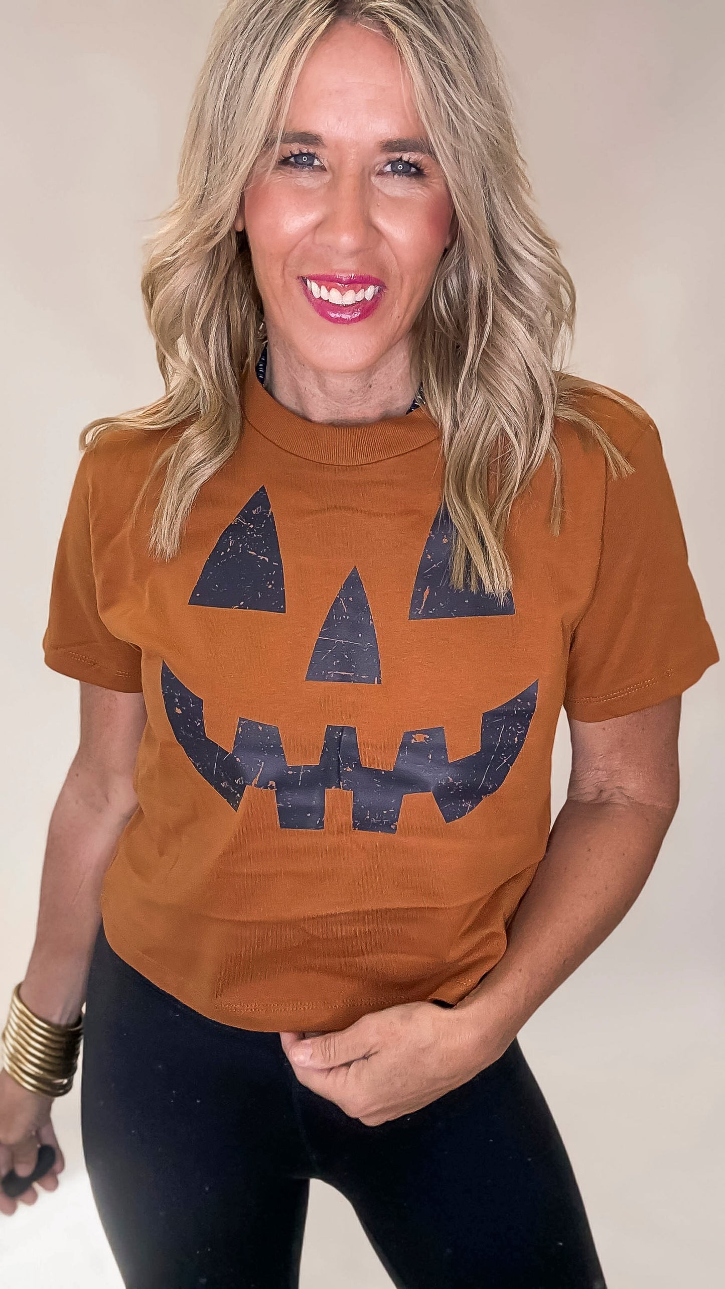 Woman wearing a antique gold t-shirt with a jack-o'-lantern face design on a beige background
