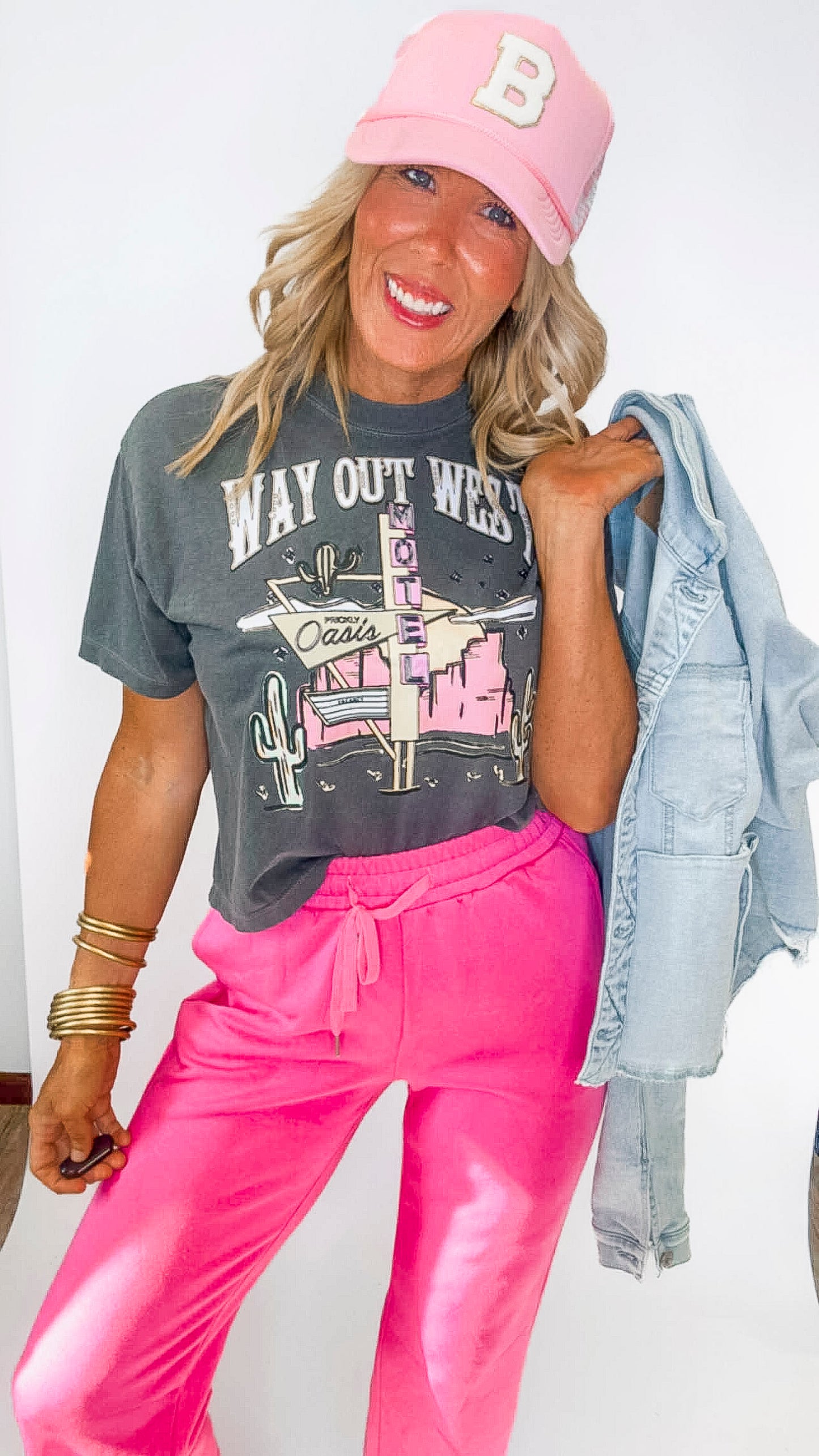 Woman wearing a gray 'Way Out West' t-shirt, pink pants, and a pink cap with a 'B' logo.