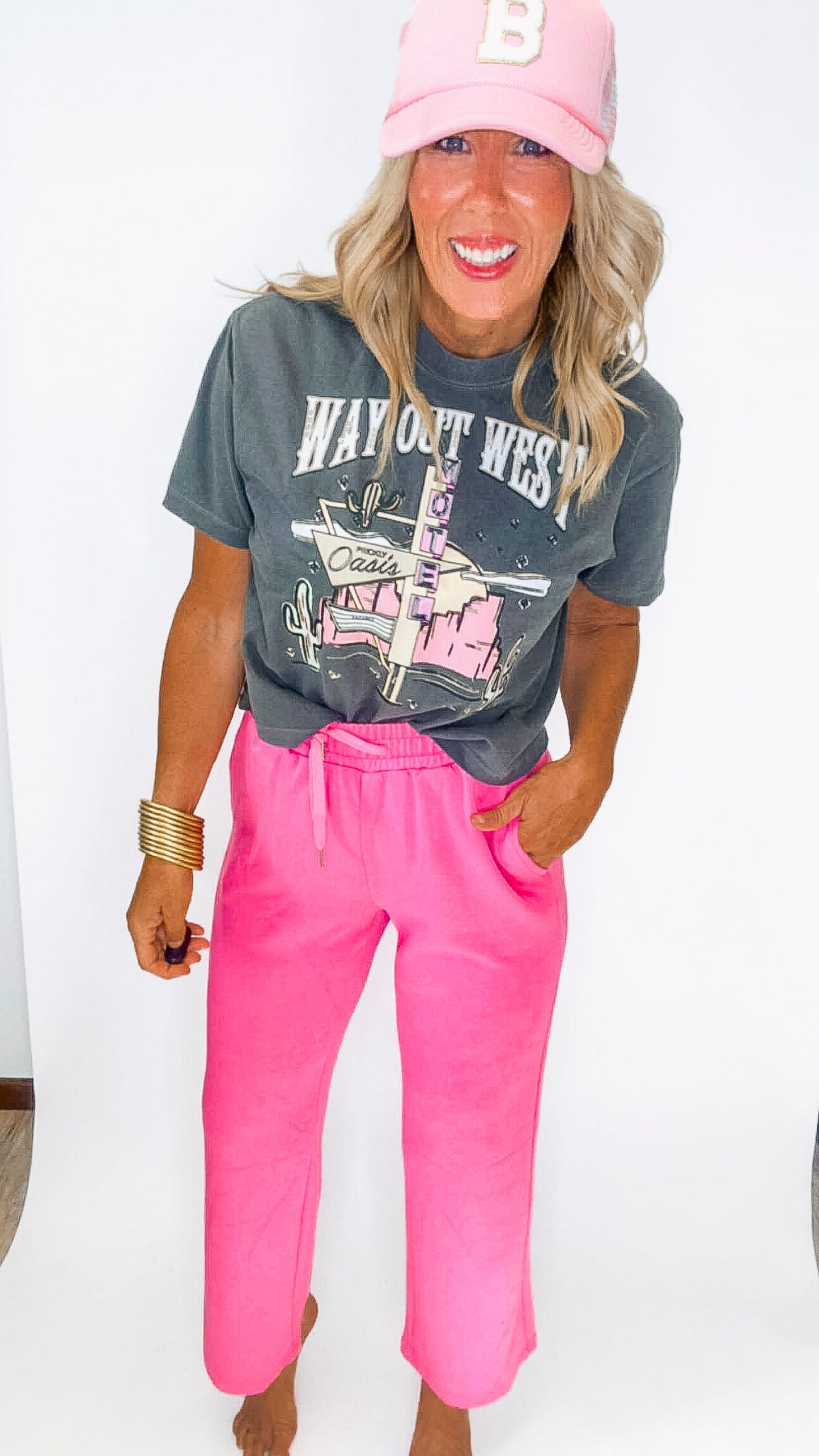 Person wearing a gray 'Way Out West' t-shirt, pink pants, and a pink cap on a white background