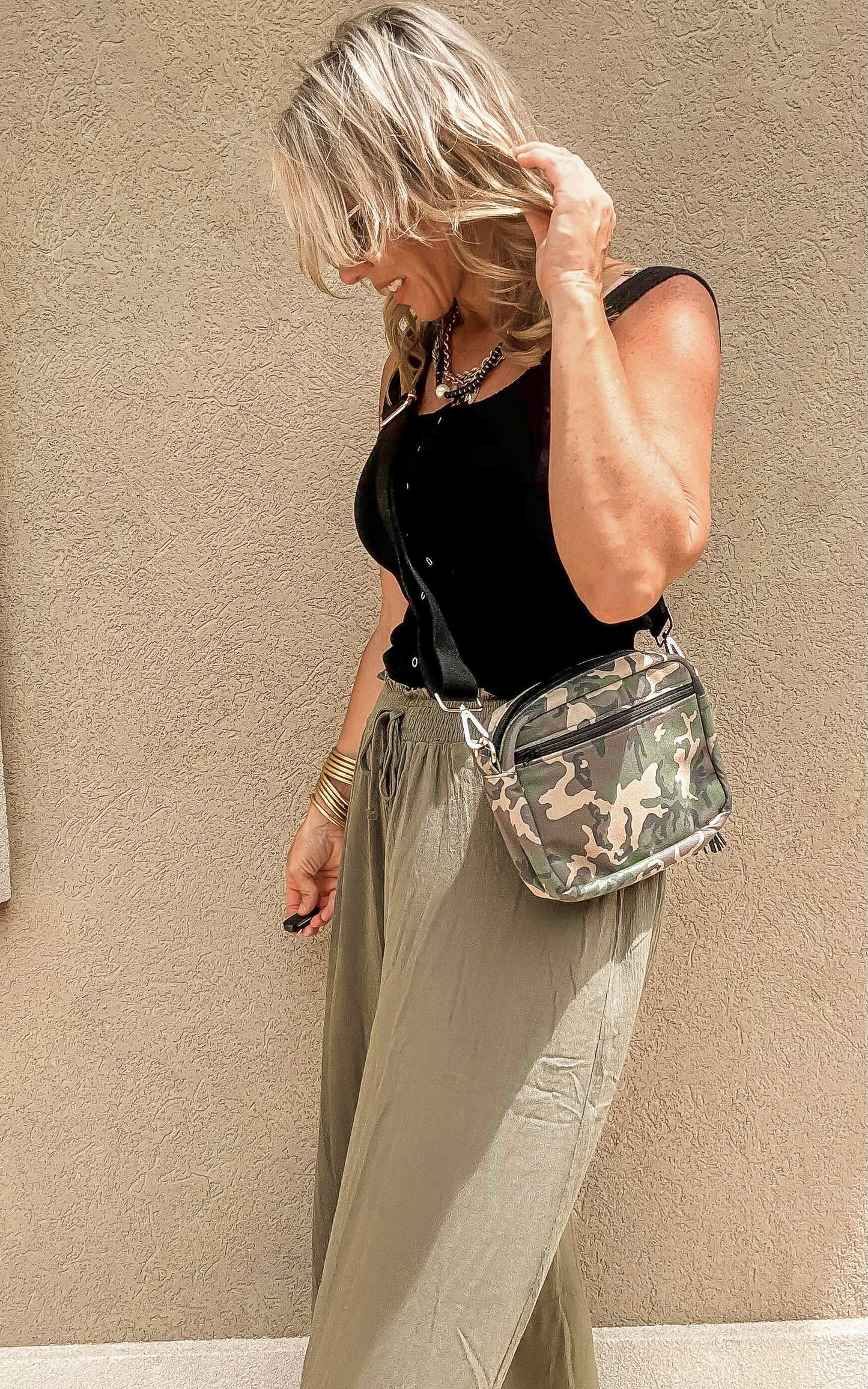 Woman wearing olive pants and a black tank holding the camo Uptown crossbody bag from the Makeup Junkie x Bad Habit collab.