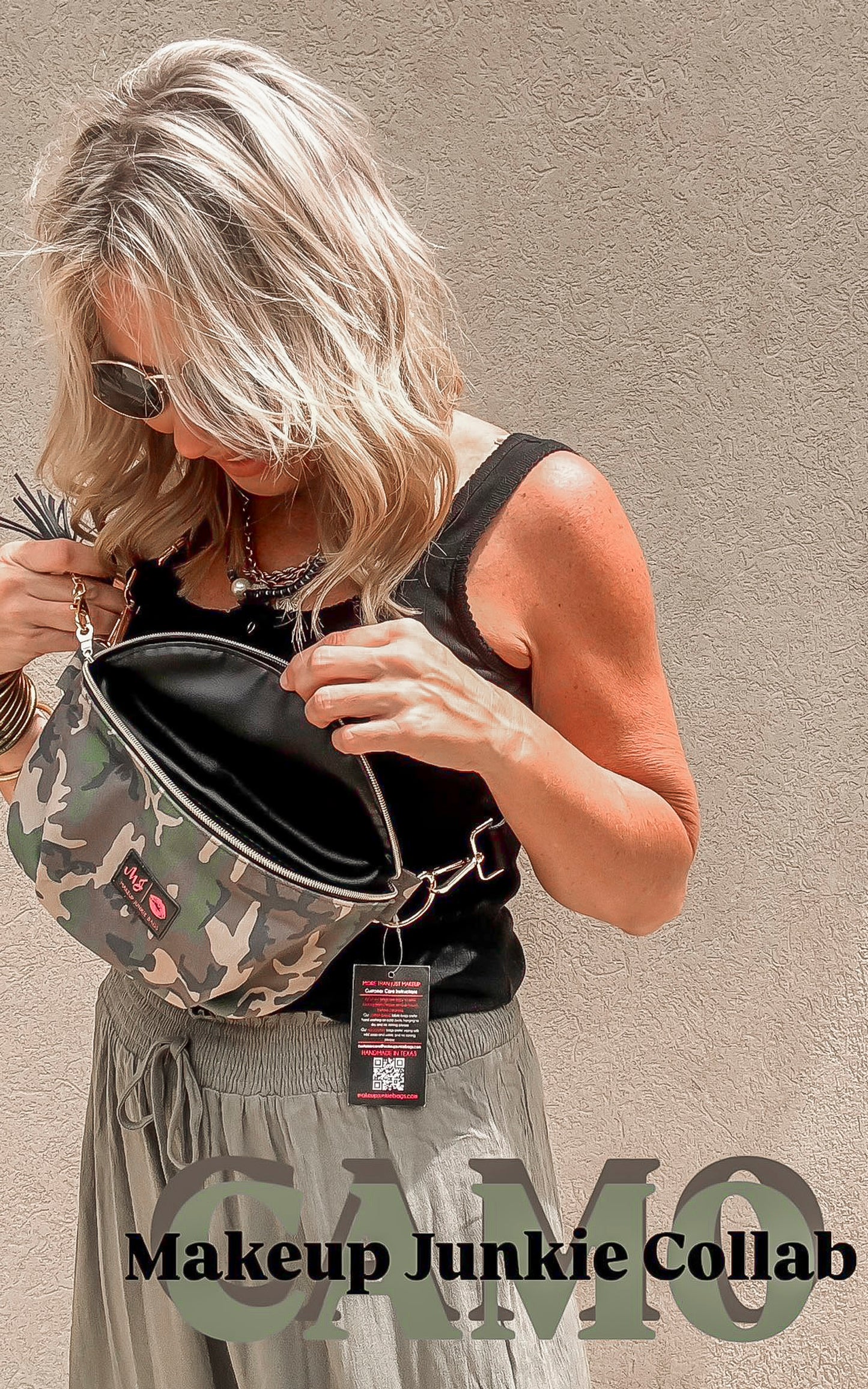 Woman holding a camo print crossbody sidekick bag by Makeup Junkie & Bad Habit, styled with a black tank and olive pants.