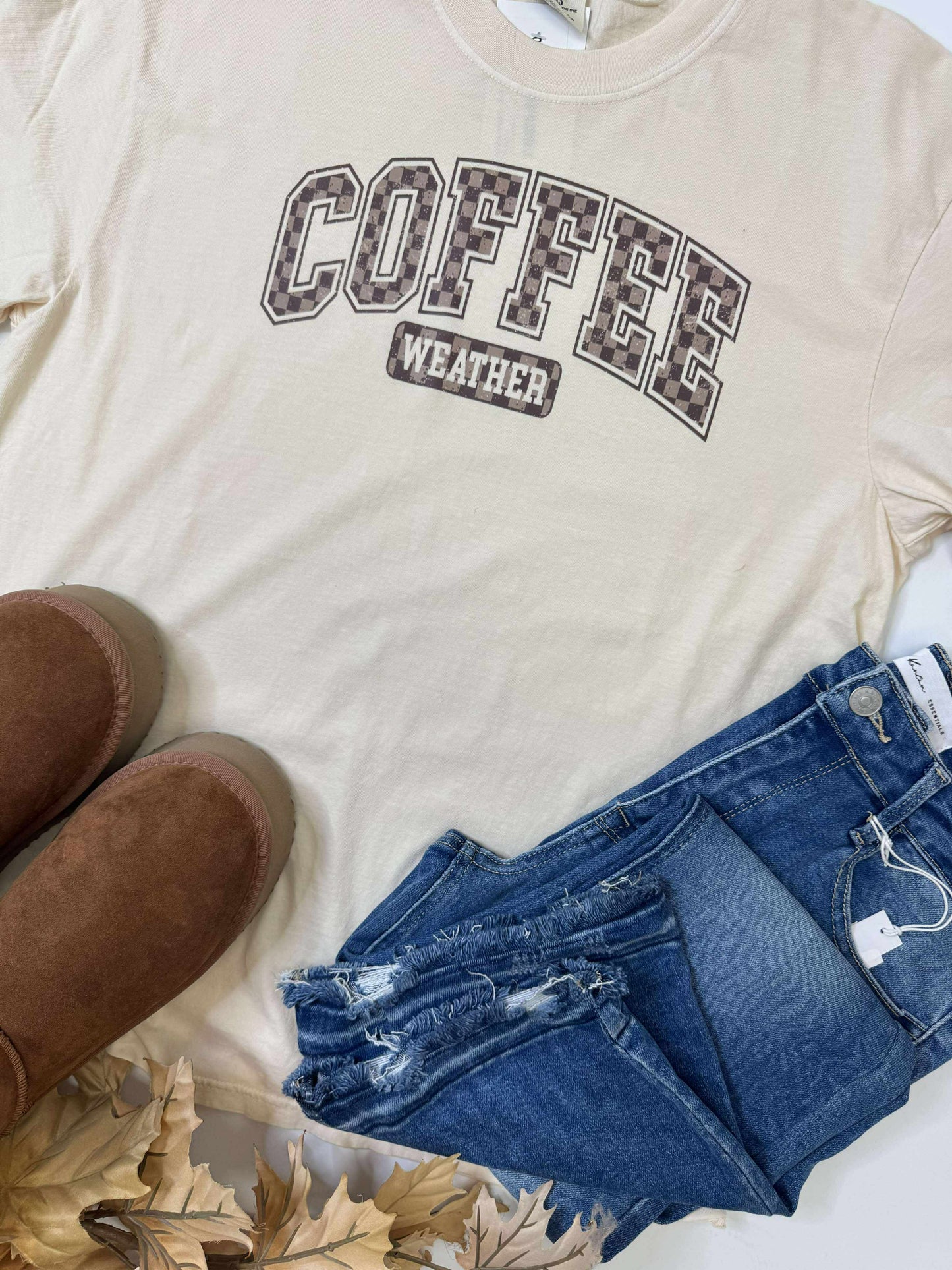 Beige t-shirt with 'COFFEE WEATHER' text, blue jeans, and brown shoes on a white surface.