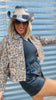 Person wearing a leopard print jacket, black top, and shorts with a cowboy hat against a corrugated metal background.