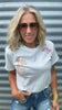 Woman wearing a white t-shirt with floral designs, sunglasses, and blue jeans against a corrugated metal background.