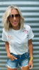 Woman wearing a white t-shirt with graphics and denim shorts against a corrugated metal background