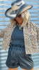 Person wearing a leopard print jacket, black top, and leather shorts with a wide-brimmed hat against a metallic background.
