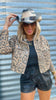 Person wearing a leopard print jacket, black shorts, and a straw hat against a corrugated metal background.