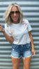 Woman wearing a white floral embroidered t-shirt and denim shorts against a corrugated metal background.