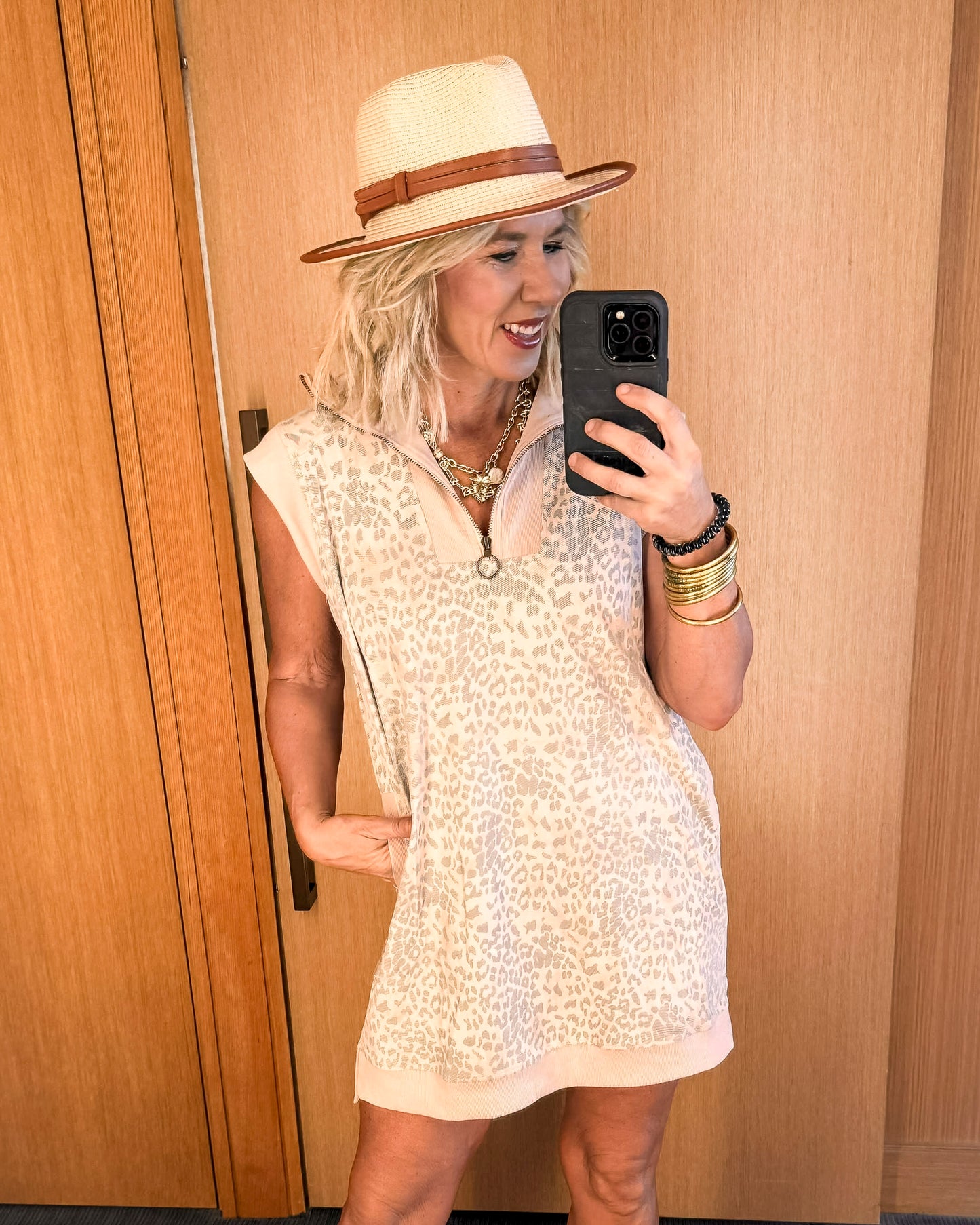 Woman taking a mirror selfie wearing a white dress and beige hat against a wooden wall.