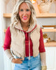 Woman wearing a beige vest over a red shirt with blue jeans, standing in a room with wooden shelves.