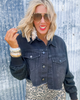 Woman wearing a denim jacket and sunglasses against a textured wall.