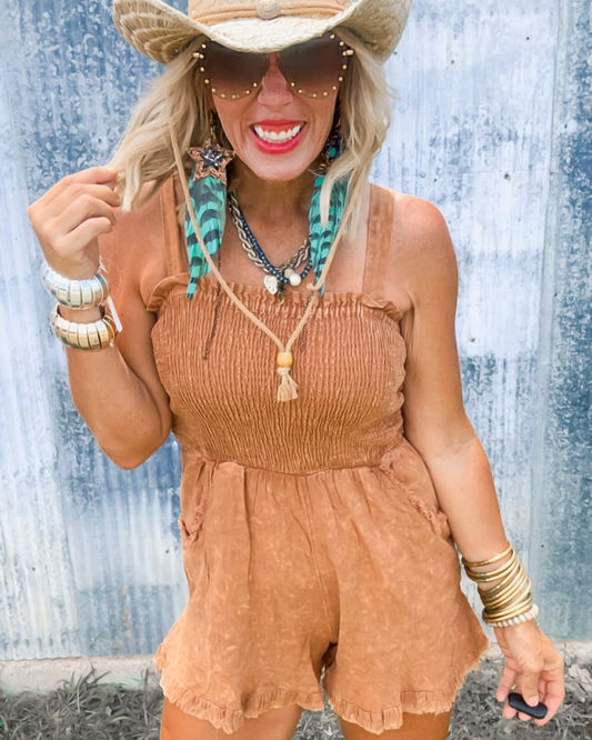 Woman wearing a tan romper with accessories against a textured blue wall.