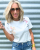 Woman wearing a white t-shirt with colorful designs and blue jeans against a metal wall.