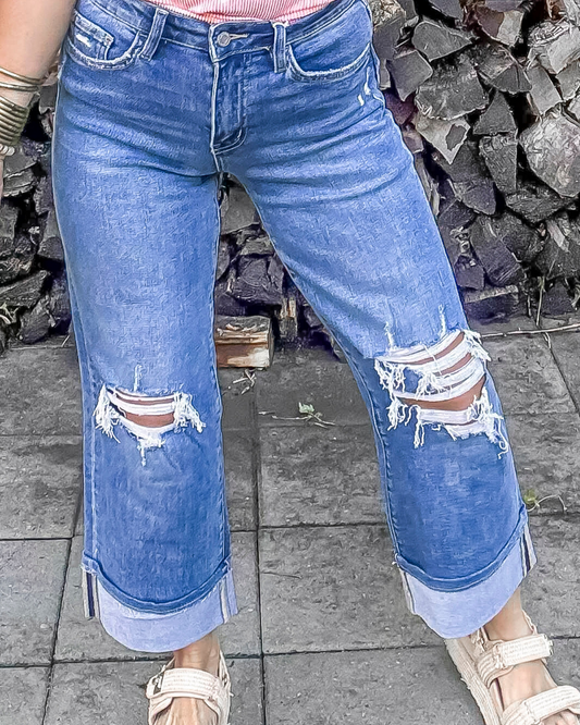 Person wearing blue ripped jeans standing on a stone pavement with a stone wall background.