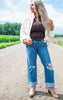 Woman standing on a dirt road wearing a white jacket, brown top, blue jeans with fish designs, and beige boots.