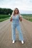Woman in a light blue jumpsuit standing on a rural road.