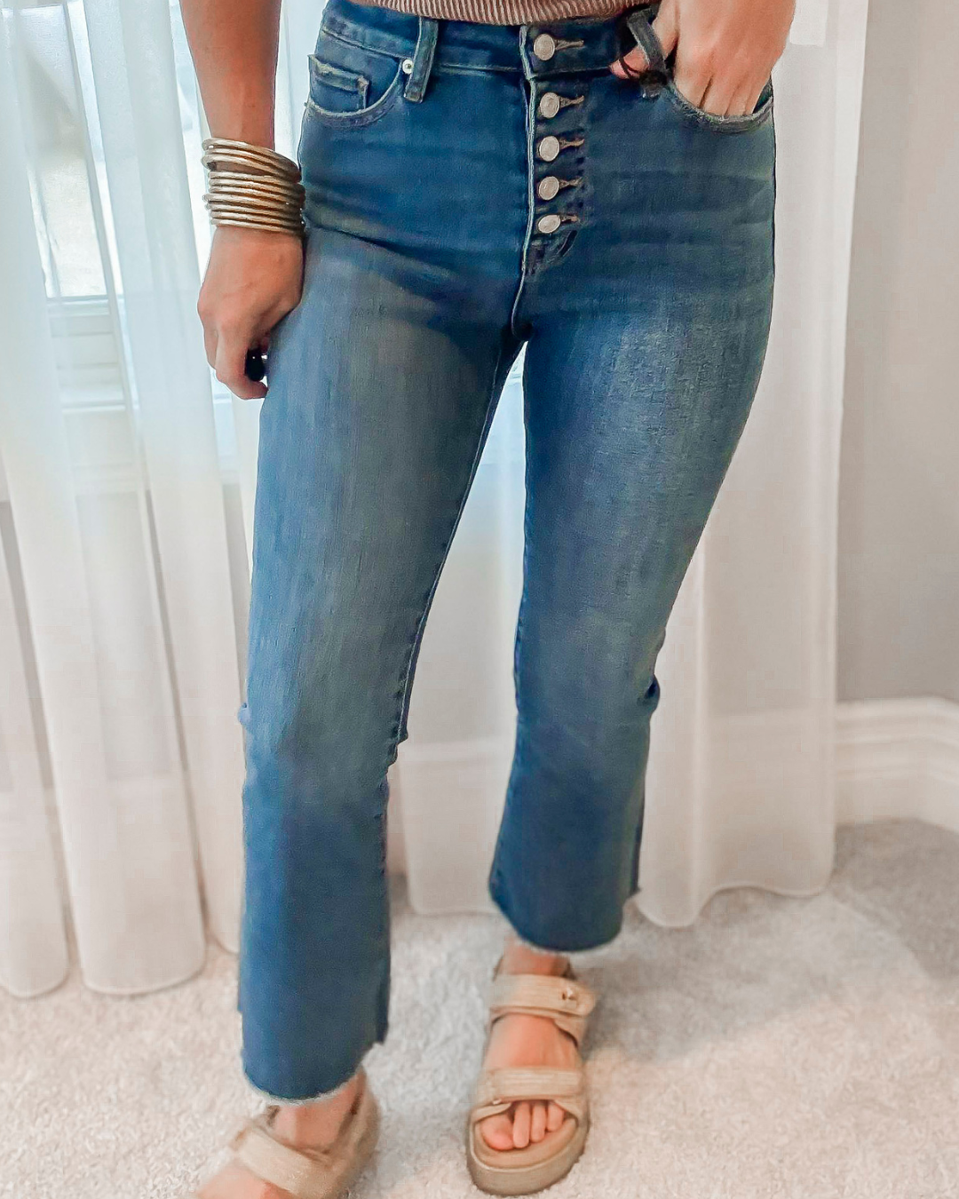 Person wearing blue jeans with a button fly, standing indoors with white curtains in the background.