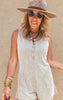 Woman in a sand-colored ribbed romper with front buttons and patch pockets, accessorized with layered necklaces and a wide-brim hat.