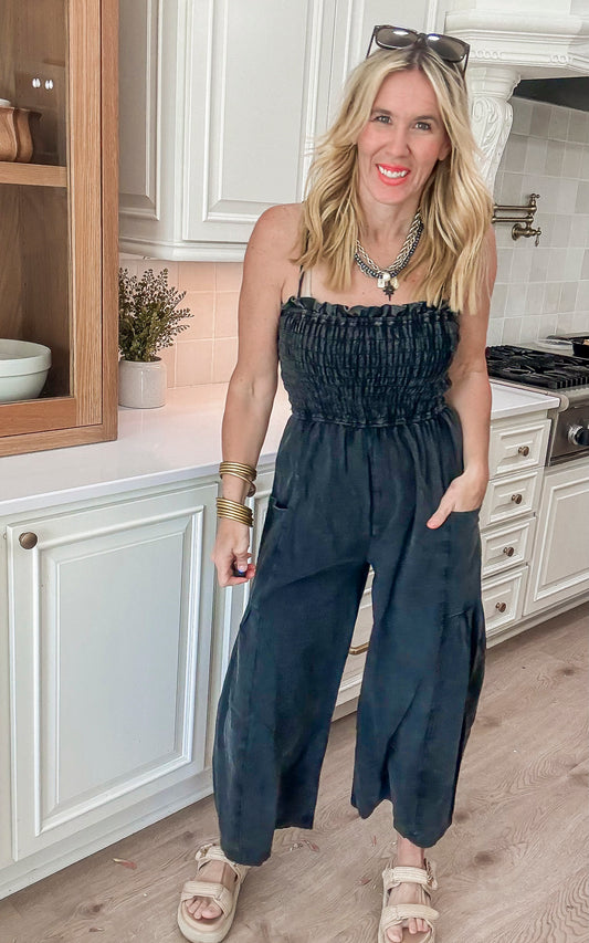 Woman wearing a black smocked strapless jumpsuit with wide legs, styled casually with sandals and layered necklaces.