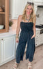 Woman wearing a black smocked strapless jumpsuit with wide legs, styled casually with sandals and layered necklaces.
