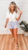 Woman in a white blouse and light blue shorts standing in a room with wooden flooring and a plant.