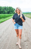 Woman standing on a dirt road in a field wearing a black shirt, denim shorts, and beige boots.
