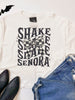 White t-shirt with 'SHAKE ME SENORA' text and bow graphic, paired with denim shorts and black shoes on a white background.