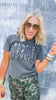 Woman wearing a gray t-shirt with text and camouflage pants against a textured wall.