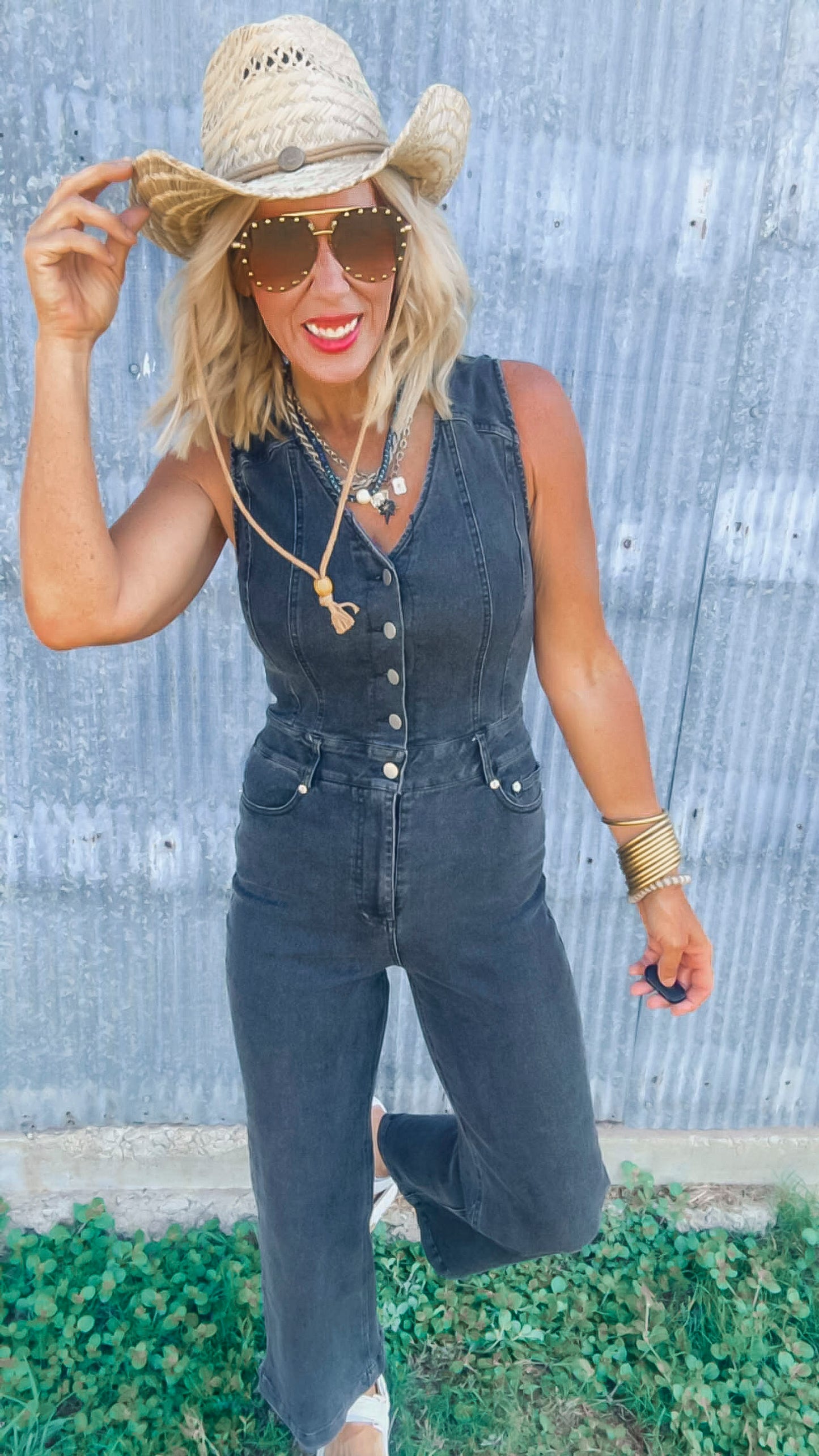 Woman wearing a denim jumpsuit with a straw hat and sunglasses against a corrugated metal background.