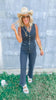 Woman wearing a denim jumpsuit and straw hat in front of a corrugated metal wall.