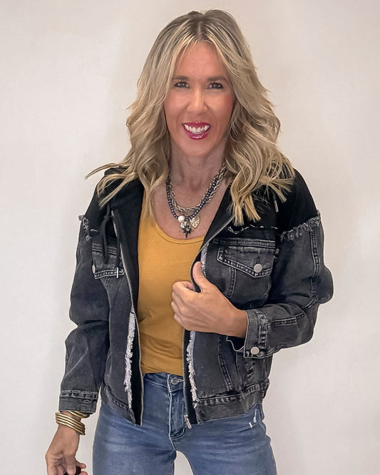 Woman wearing a black denim jacket over a yellow top and blue jeans on a plain background