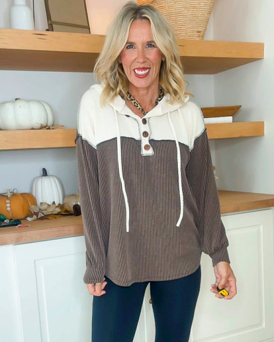 Woman wearing a brown and white long-sleeve top with a hood, standing in a kitchen.